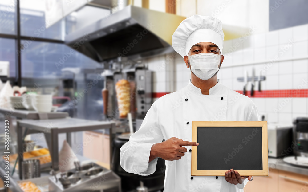 health protection, safety and pandemic concept - indian male chef cook wearing face protective mask with blank chalkboard over kebab shop kitchen background