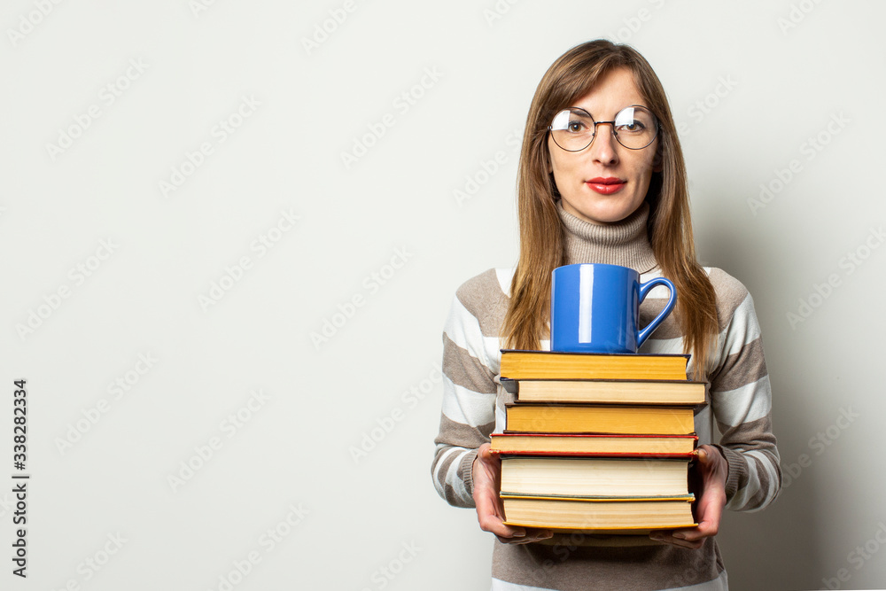 Portrait of a young friendly woman in a sweater and glasses holding a stack of books and a cup with coffee in an isolated light background. Emotional face. Education concept, exam preparation Portrait of a young friendly woman in a sweater and glasses holding a stack of books and a cup with coffee in an isolated light background. Emotional face. Education concept, exam preparation