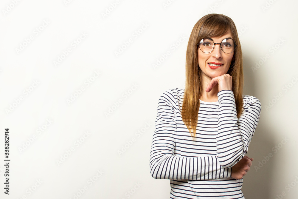 Portrait of a young friendly woman in a long-sleeve sweater and glasses with a smile, holds her hand under her chin on an isolated light background. Emotional face. Gesture to think, dream Portrait of a young friendly woman in a long-sleeve sweater and glasses with a smile, holds her hand under her chin on an isolated light background. Emotional face. Gesture to think, dream
