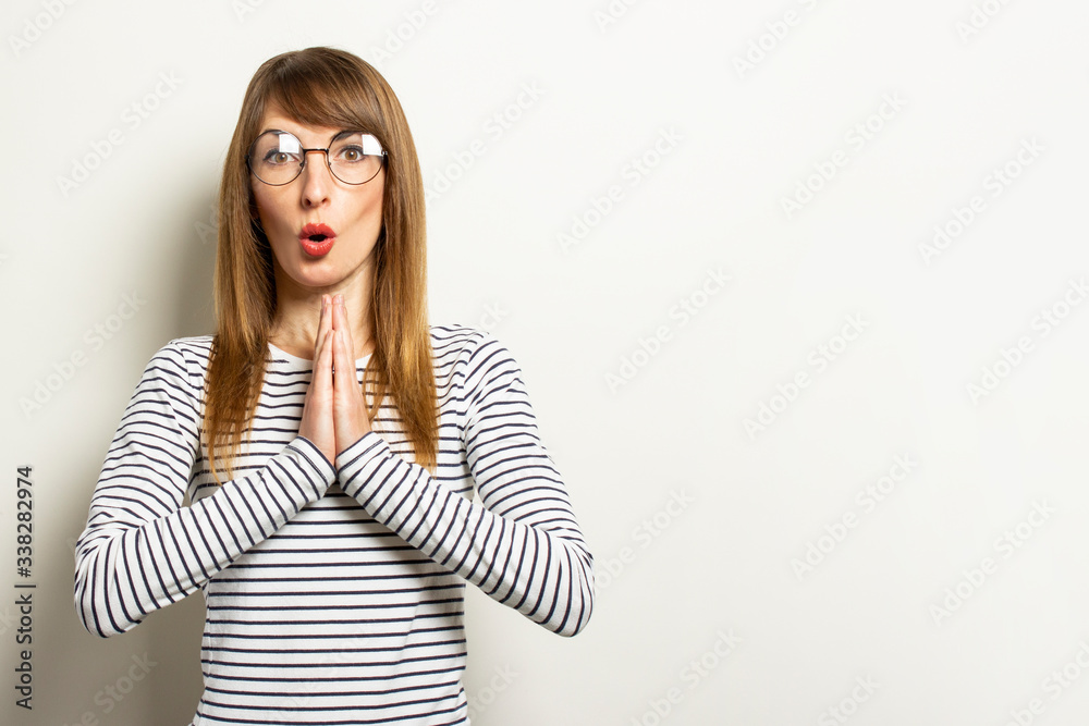 Young friendly woman in a long-sleeve sweater and glasses with a surprised face and folded arms for prayer on an isolated light background. Emotional face. Gesture of meditation, prayer, relaxation Young friendly woman in a long-sleeve sweater and glasses with a surprised face and folded arms for prayer on an isolated light background. Emotional face. Gesture of meditation, prayer, relaxation
