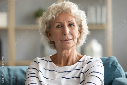 Head shot portrait close up beautiful aged mature woman with grey curly hair sitting on cozy couch, posing for photo at home, attractive older senior female looking at camera, natural old beauty