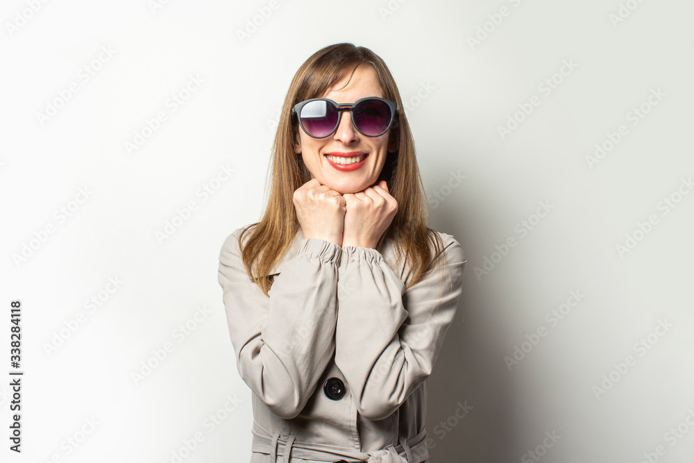 Portrait of a young friendly woman in a classic jacket and sunglasses holding her hands under her chin and smiling on an isolated light background. Emotional face. Classic style concept, french woman Portrait of a young friendly woman in a classic jacket and sunglasses holding her hands under her chin and smiling on an isolated light background. Emotional face. Classic style concept, french woman