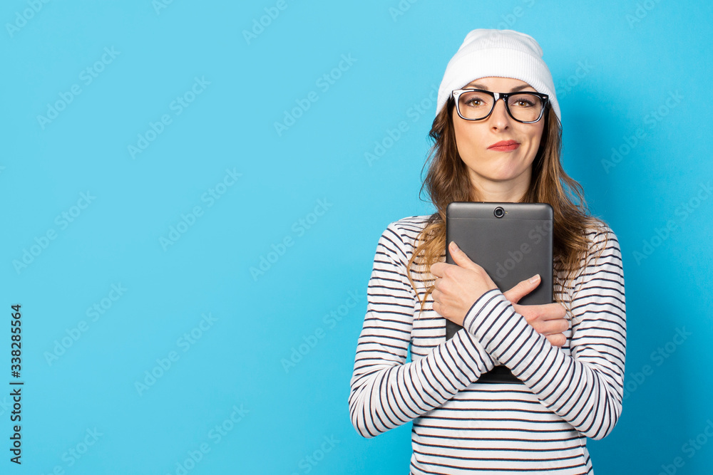 Portrait of a young friendly woman with an embarrassed face in a hat, glasses and a T-shirt holding a tablet in her hands on an isolated blue background. Emotional face Portrait of a young friendly woman with an embarrassed face in a hat, glasses and a T-shirt holding a tablet in her hands on an isolated blue background. Emotional face