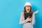 Portrait of a young friendly woman with a surprised face in a hat, glasses and a T-shirt holding a tablet in her hands and looking at it on an isolated blue background. Emotional face