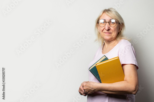 Portrait of an old friendly woman with smile in a casual t-shirt and glasses holds a stack of books on an isolated light background. Emotional face. Concept book club, leisure, education