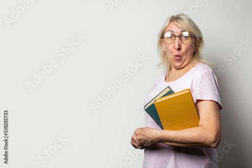 Portrait of an old friendly woman with a surprised face in a casual t-shirt and glasses holds a stack of books on an isolated light background. Emotional face. Concept book club, leisure, education