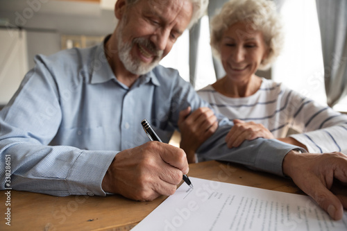 Close up satisfied older couple signing documents, making purchasing or investment deal, smiling mature husband and wife holding hands, putting signature on contract, buying house or taking loan