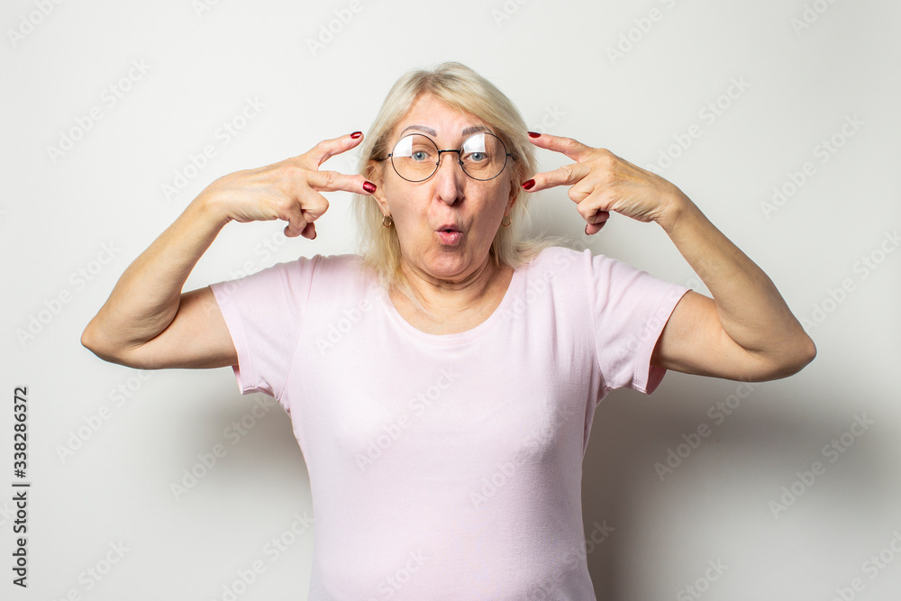 Portrait of an old friendly woman with a surprised face in a casual t-shirt and glasses makes a welcome gesture on an isolated light background Portrait of an old friendly woman with a surprised face in a casual t-shirt and glasses makes a welcome gesture on an isolated light background