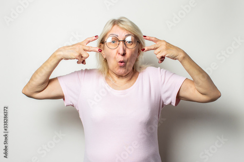 Portrait of an old friendly woman with a surprised face in a casual t-shirt and glasses makes a welcome gesture on an isolated light background