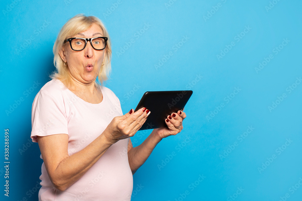 Portrait of an old friendly woman with a surprised face in glasses and an casual t-shirt holding a tablet in her hands on an isolated blue background. Emotional face Portrait of an old friendly woman with a surprised face in glasses and an casual t-shirt holding a tablet in her hands on an isolated blue background. Emotional face