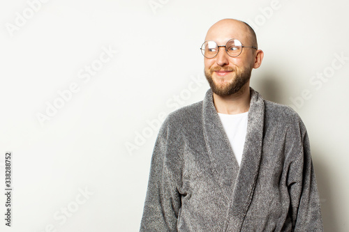 Portrait of a young bald man with a beard in a bathrobe and glasses looks away on an isolated light background. Emotional face