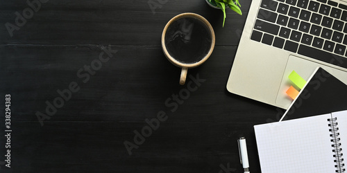 Top view image of computer laptop putting on black working desk and surrounded by notebook, diary, potted plant, coffee cup and pen. Orderly workspace concept.