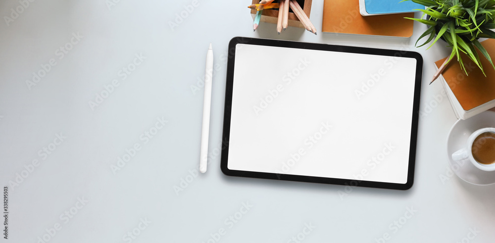 Top view image of computer tablet with white blank screen putting on white working desk and surrounded by stylus pen, potted plant, notebook, diary, coffee cup, pencils and pencil holder. Top view image of computer tablet with white blank screen putting on white working desk and surrounded by stylus pen, potted plant, notebook, diary, coffee cup, pencils and pencil holder.