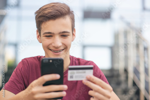 Teenage boy with a credit card and mobile phone makes purchasing outdoors. Happy young man is using smartphone and bank card for online shopping. Handsome smiling guy holds bank card and cell phone