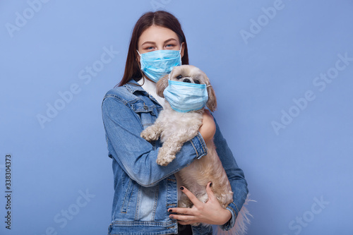 Horizontal shot of young attractive lady with dark hair holding her little pet with protective mask, female wearing denim jacket and flumask, looks at camera, girl tries to avoid coronavirus infection photo