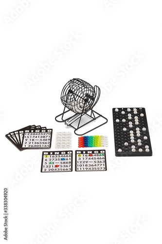 Subject shot of a bingo game on the white backdrop. The game set consists of a black cage, bingo cards, colored bingo chips, white bingo balls and a tray for balls. 