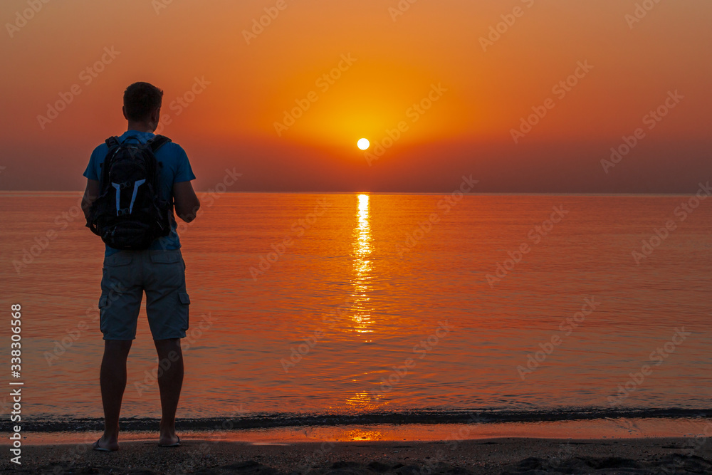 A man with a backpack stands on the beach and watches the sun rise.