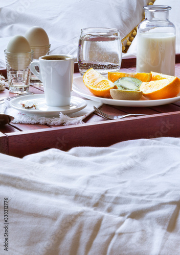 Breakfast in bed on wooden tray with fruits, milk, eggs and coffe. Window light, copy space