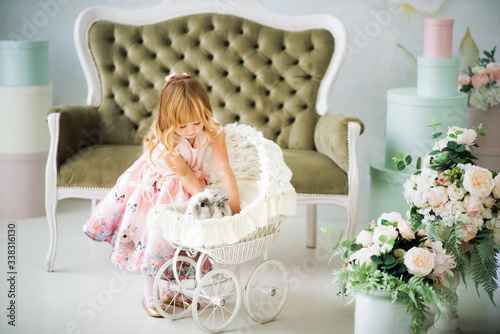 Little pretty smiling blonde 3 years old girl in pink dress putting gray and white furry rabbit in white baby carriage. Child and bunny. Spring easter holidays. Friendship between child and animal