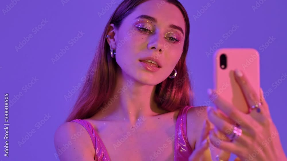 A cheerful woman with fashion glitter makeup is making selfie photos isolated over violet neon background A cheerful woman with fashion glitter makeup is making selfie photos isolated over violet neon background