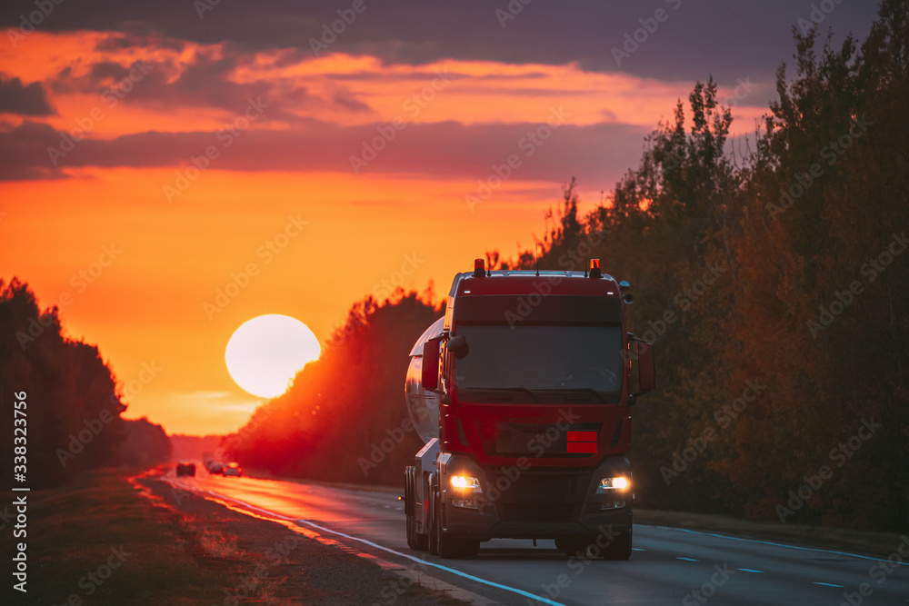 Red Truck Or Tractor Unit, Prime Mover, Traction Unit In Motion On Road, Freeway. Asphalt Motorway Highway Against Background Of Big Sunset Sun. Business Transportation And Trucking Industry.