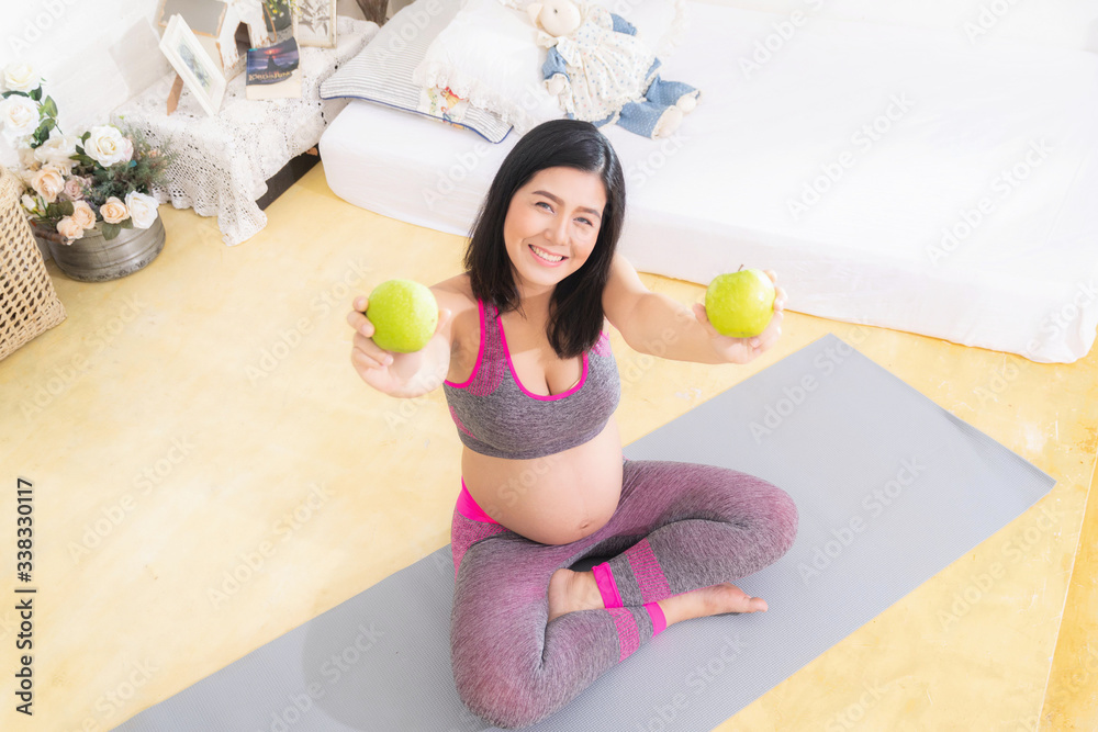 Pregnant woman wearing pink exercise clothing. Care about unborn child. Happy pregnant woman eating natural green apple, sitting at bedroom. Pregnant woman wearing pink exercise clothing. Care about unborn child. Happy pregnant woman eating natural green apple, sitting at bedroom.