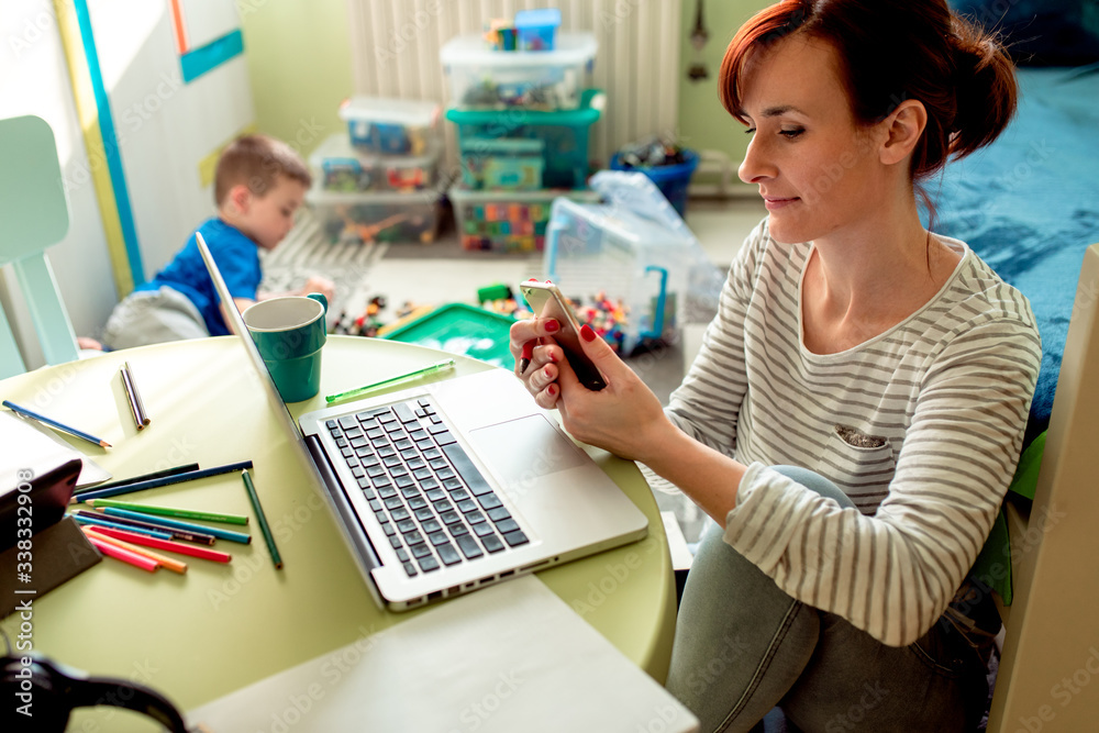 Mother working remotely on laptop while taking care of her son playing with toys in his room. Mother working remotely on laptop while taking care of her son playing with toys in his room.