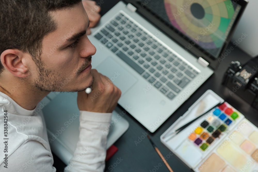 Top view of designer thinking on the workplace hold the electronic work pen. Desktop with laptop, camera, watercolors on a dark table. Top view of designer thinking on the workplace hold the electronic work pen. Desktop with laptop, camera, watercolors on a dark table.