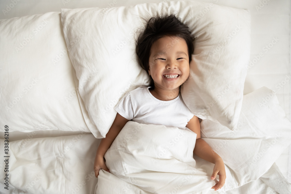 Top view in bed fresh sheets comfortable mattress soft pillow white bedding lies small asian girl has wide charming toothy smile looks at camera, kid woke up, enough healthy sleep, feels active lively - obrazy, fototapety, plakaty 