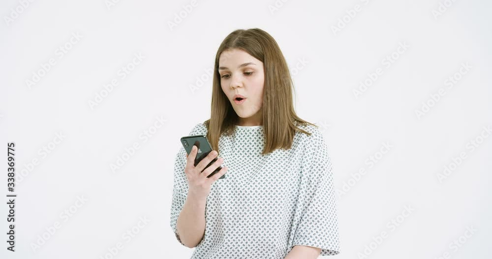 Camera zooming in at beutiful female Caucasian medical patient texting massage on mobile phone on white wall background. Smiled pretty young girl scrolling and tapping on smartphone in hospital. Camera zooming in at beutiful female Caucasian medical patient texting massage on mobile phone on white wall background. Smiled pretty young girl scrolling and tapping on smartphone in hospital.