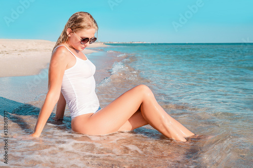 Beautiful girl sunbathing on a sandy tropical beach. Fashion young female lying on back on sand with white swimsuit. Tanned blonde woman relaxing