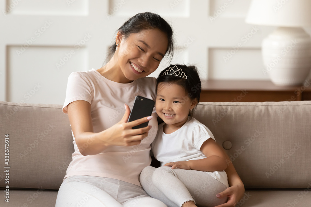 Cheerful Asian mother cuddles little sweet daughter holding smartphone making video call talk to relatives, taking selfie photography for memory, having fun at home use modern application tech concept