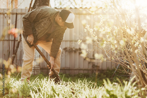 Adult man digging ground on backyard with shovel. Working with both hands. Agricultural life in spring. Trees have blossom. Sunny day outside.