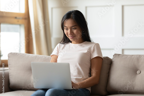 Asian young woman hold on lap notebook while typing e-mail seated on couch in living room alone. Social network media active user, do remote work, shopper makes goods services purchase on-line concept