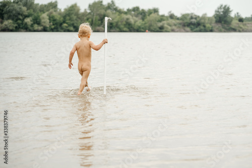 Theme summer outdoor activities near the river on the city beach in Russia. Little funny naked baby boy walks on the water on the river bank, jumping a splash of water sandy beach summer time.