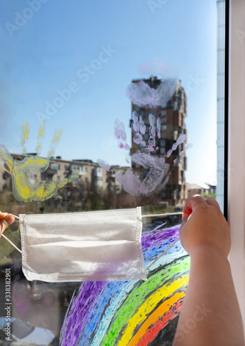 children's hands hold a protective mask near the window with colored palm prints and a rainbow