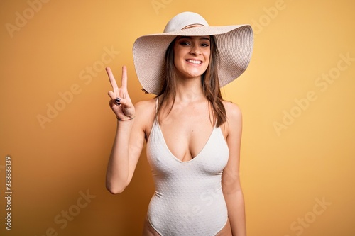 Young beautiful brunette woman on vacation wearing swimsuit and summer hat showing and pointing up with fingers number two while smiling confident and happy.