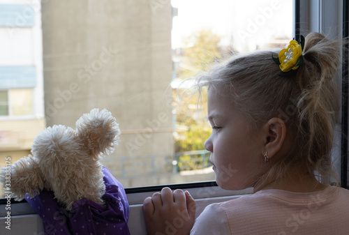 self-isolation. quarantine. a small blonde girl with a stuffed toy looks out the window at the street