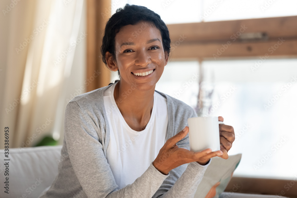 Portrait of happy and smiling young african american girl, sit and rest on sofa in living room, hold cup in her hands, drink and enjoy tea or coffee, lazy relaxation day, weekend at home Portrait of happy and smiling young african american girl, sit and rest on sofa in living room, hold cup in her hands, drink and enjoy tea or coffee, lazy relaxation day, weekend at home