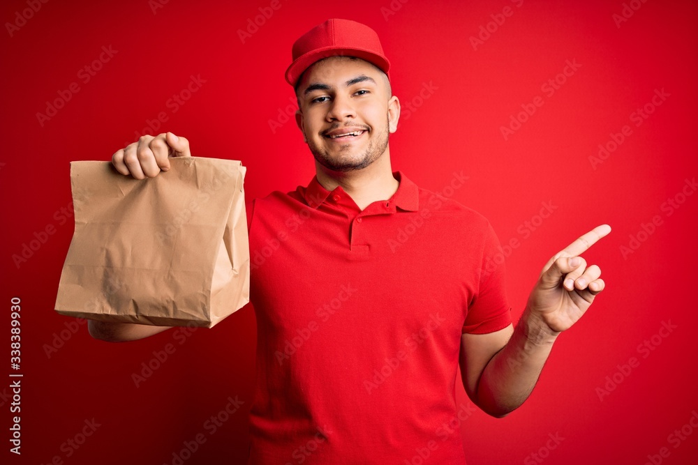 Young handsome delivery man holding paper bag with takeaway food over red background very happy pointing with hand and finger to the side