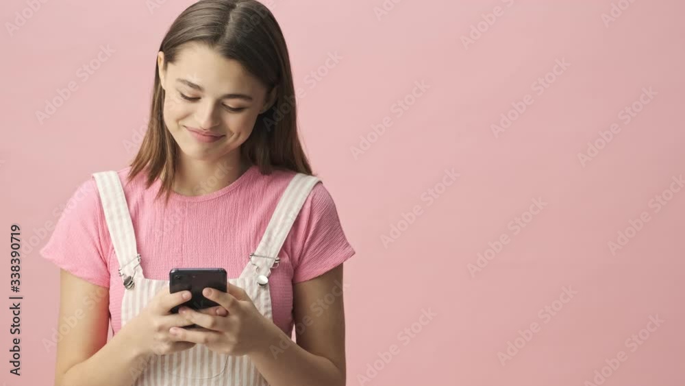 Happy pretty brunette woman in overalls using smartphone over pink background Happy pretty brunette woman in overalls using smartphone over pink background