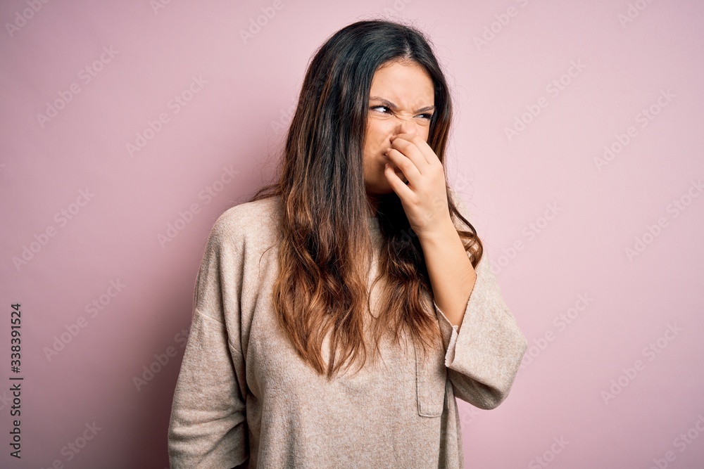 Young beautiful brunette woman wearing casual sweater standing over pink background smelling something stinky and disgusting, intolerable smell, holding breath with fingers on nose. Bad smell - obrazy, fototapety, plakaty 