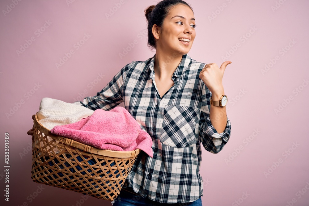 Young beautiful brunette woman doing chores holding wicker basket with clothes pointing and showing with thumb up to the side with happy face smiling Young beautiful brunette woman doing chores holding wicker basket with clothes pointing and showing with thumb up to the side with happy face smiling