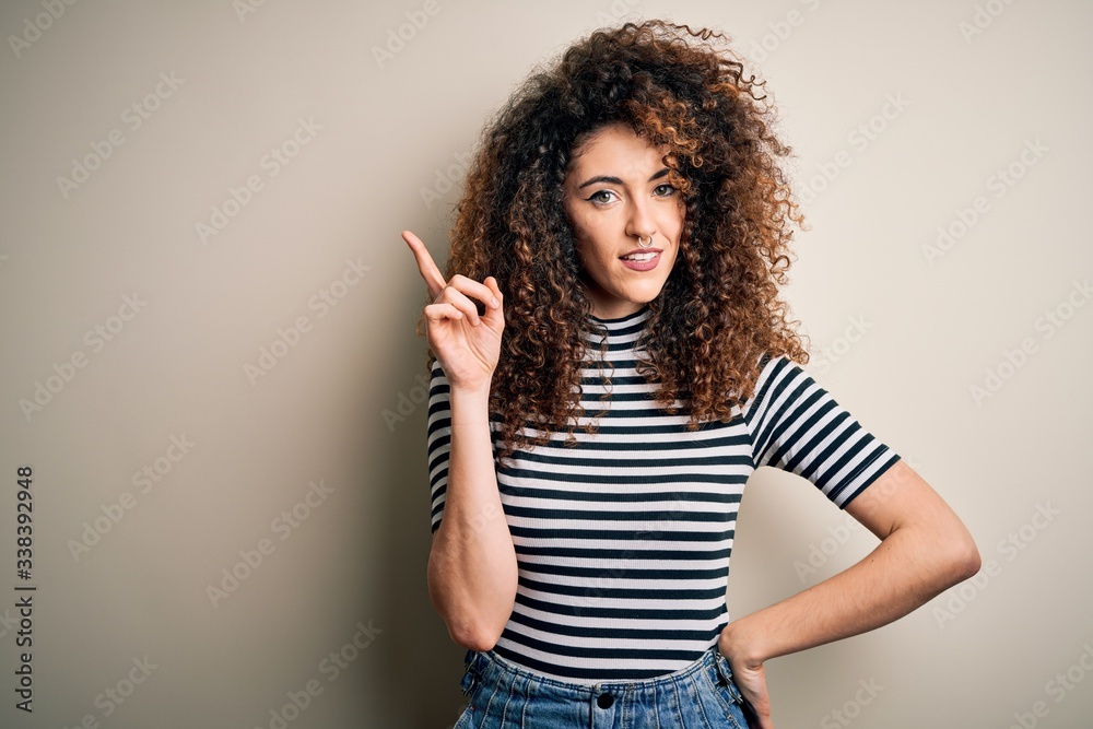 Young beautiful woman with curly hair and piercing wearing casual striped t-shirt with a big smile on face, pointing with hand and finger to the side looking at the camera. Young beautiful woman with curly hair and piercing wearing casual striped t-shirt with a big smile on face, pointing with hand and finger to the side looking at the camera.