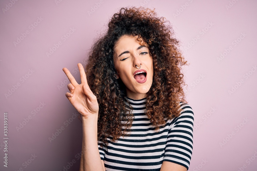 Young beautiful woman with curly hair and piercing wearing casual striped t-shirt smiling with happy face winking at the camera doing victory sign. Number two. Young beautiful woman with curly hair and piercing wearing casual striped t-shirt smiling with happy face winking at the camera doing victory sign. Number two.
