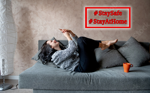 Stayathome staysafe hashtag sign on young woman smiling and using mobile phone at home for coronavirus quarantine photo