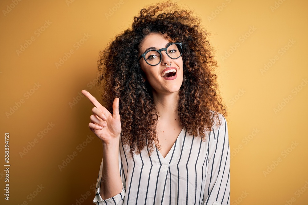 Young beautiful woman with curly hair and piercing wearing striped shirt and glasses pointing finger up with successful idea. Exited and happy. Number one. Young beautiful woman with curly hair and piercing wearing striped shirt and glasses pointing finger up with successful idea. Exited and happy. Number one.