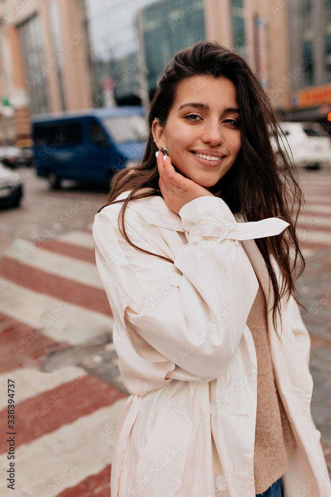 Fototapeta premium Outside portrait of attractive woman with dark hair wearing white coat walking on the street with charming smile in good day