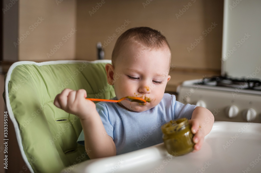 The first bait. Baby eats at the high chair in the kitchen food for babies. Good nutrition. Baby in a blue bodysuit eats broccoli.the boy eats on his own The first bait. Baby eats at the high chair in the kitchen food for babies. Good nutrition. Baby in a blue bodysuit eats broccoli.the boy eats on his own