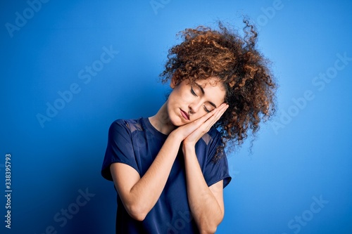 Young beautiful woman with curly hair and piercing wearing casual blue t-shirt sleeping tired dreaming and posing with hands together while smiling with closed eyes.